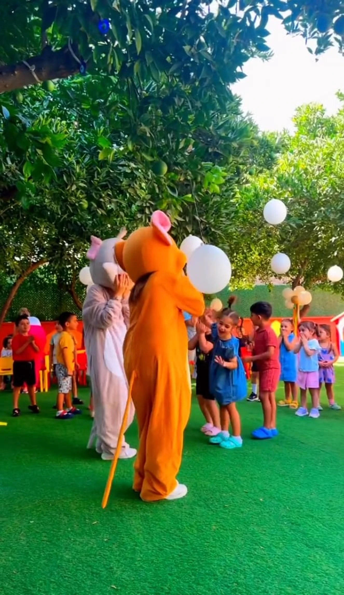 Portakal ağaçlarıyla çevrili bahçede maskot kostümlü karakterlerle oyun oynayan çocuklar